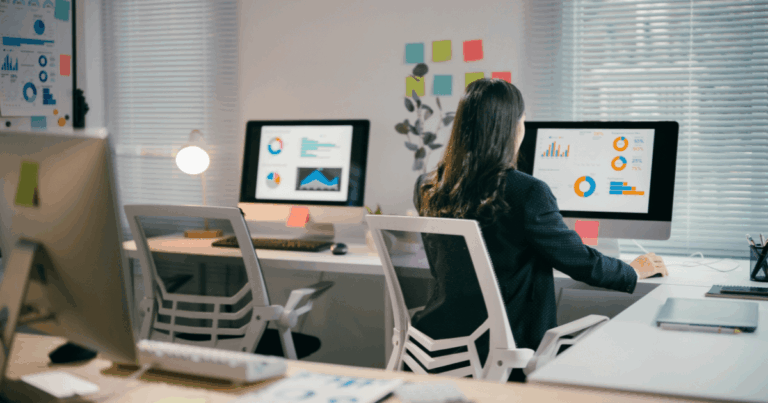 Profissional trabalhando em escritório moderno, analisando indicadores de gestão em dashboards com gráficos e métricas exibidos em telas de computador.