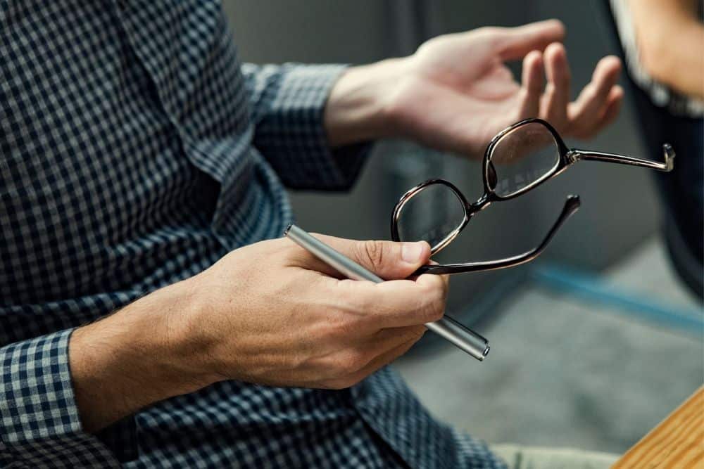 Recorte de mãos masculinas gesticulando e segurando um óculos em uma reunião sobre preço de venda.