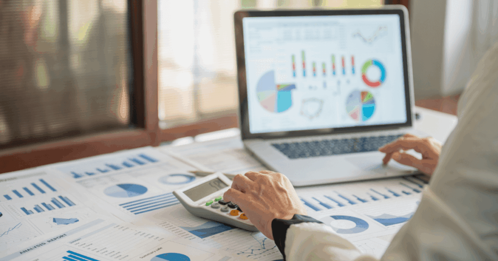 Imagem mostrando uma pessoa trabalhando com gráficos e dados de retenção de clientes. A pessoa está utilizando um computador portátil e uma calculadora, enquanto observa gráficos de barras, pizza e linhas em relatórios impressos sobre a mesa.