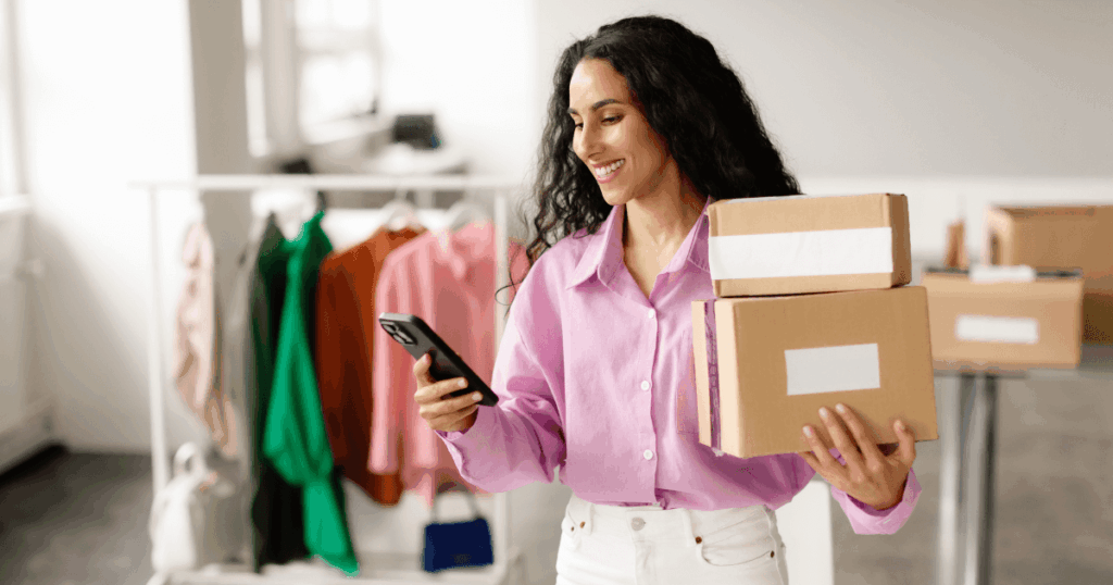 Mulher sorrindo segura caixas de envio e usa o celular em um ambiente com roupas ao fundo, representando uma vendedora organizando pedidos em um marketplace.