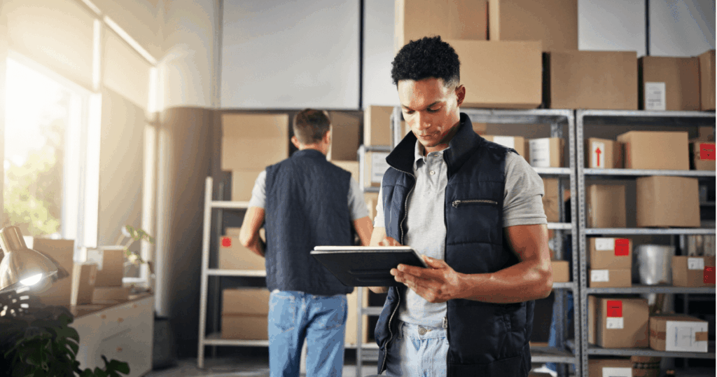 Funcionários em um armazém organizando estoques e utilizando tecnologia para controle, representando a gestão eficiente de processos que impactam diretamente os custos logísticos.