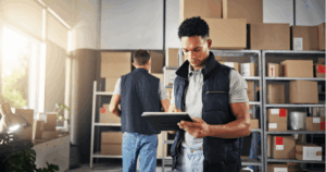Funcionários em um armazém organizando estoques e utilizando tecnologia para controle, representando a gestão eficiente de processos que impactam diretamente os custos logísticos.