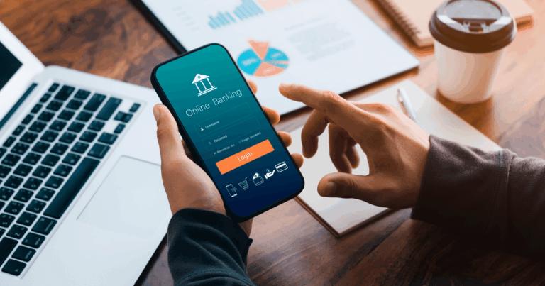 Pessoa acessando aplicativo de operações bancárias no celular, com gráficos financeiros e laptop sobre a mesa, representando gestão digital de contas e transações financeiras.