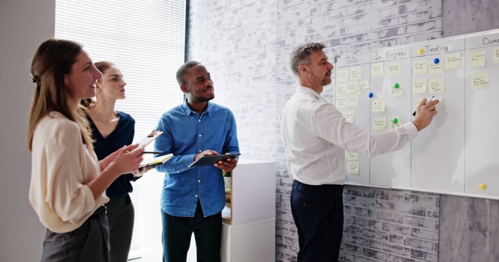 A imagem descreve 4 pessoas de um time reunidos. Eles utilizam de um quadro com post-its para representar o método Kanban para organização das tarefas.