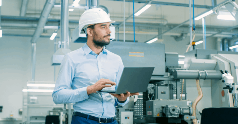 Engenheiro de produção com capacete utilizando notebook em chão de fábrica, representando o planejamento e controle da produção (PCP) na indústria.