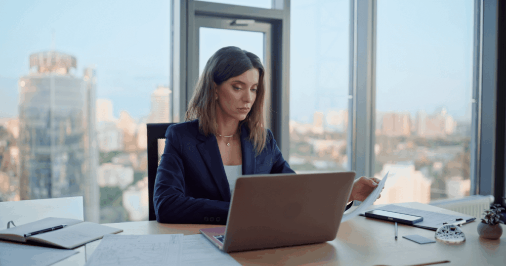 Executiva analisando documentos e trabalhando no laptop em escritório corporativo, representando gestão de contratos e acompanhamento de SLA.