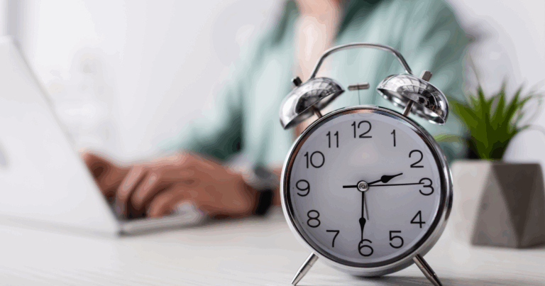 Relógio analógico marcando o horário em primeiro plano, com uma pessoa ao fundo digitando em um notebook. Representação do intervalo interjornada, destacando a importância do tempo de descanso entre turnos de trabalho para a produtividade e bem-estar do profissional.