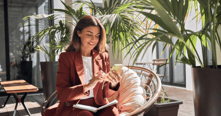 Profissional sorridente consulta dados financeiros no celular, sentada em ambiente moderno com plantas, representando praticidade e conectividade proporcionadas pelo open finance