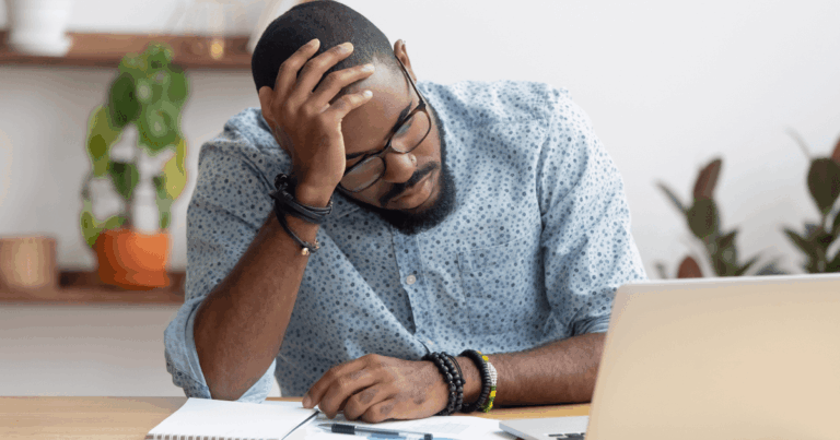 Homem com expressão de cansaço apoiando a cabeça na mão, sentado em frente ao notebook com documentos espalhados sobre a mesa, simbolizando os impactos dos riscos psicossociais no ambiente de trabalho, como estresse e esgotamento mental.