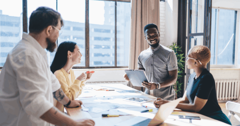 Equipe em reunião de trabalho discutindo estratégias de comunicação interna, com foco em colaboração e planejamento de ações. A imagem mostra um homem de camisa cinza apresentando ideias para colegas ao redor de uma mesa de trabalho, enquanto todos observam atentamente e interagem.