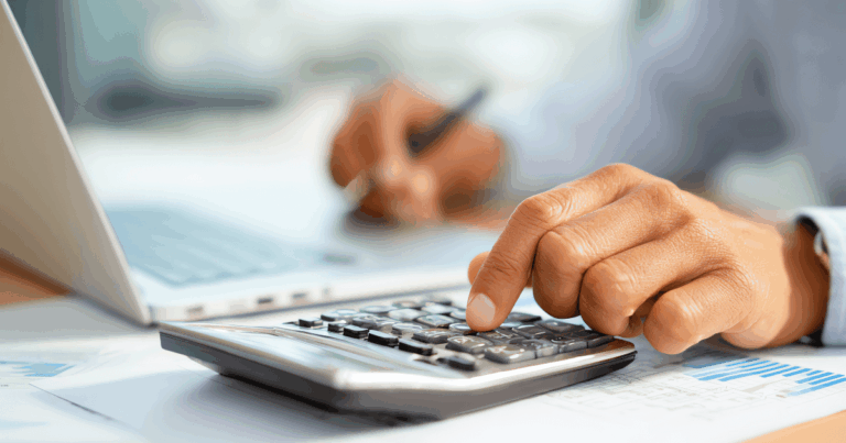 Pessoa utilizando uma calculadora e um laptop, com foco nas mãos, realizando cálculos financeiros relacionados a despesas fixas e variáveis.