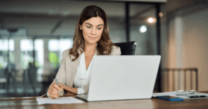 Mulher trabalhando em um escritório, olhando para o computador enquanto faz anotações, representando o ambiente de gestão de encargos trabalhistas.