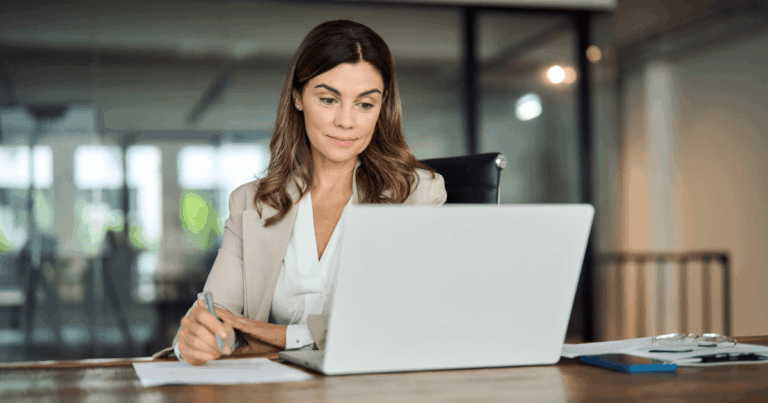 Mulher trabalhando em um escritório, olhando para o computador enquanto faz anotações, representando o ambiente de gestão de encargos trabalhistas.
