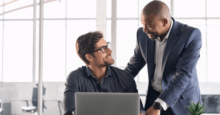 Dois profissionais em um escritório sorrindo enquanto conversam sobre um trabalho no notebook, representando colaboração e troca de experiências na jornada do colaborador.