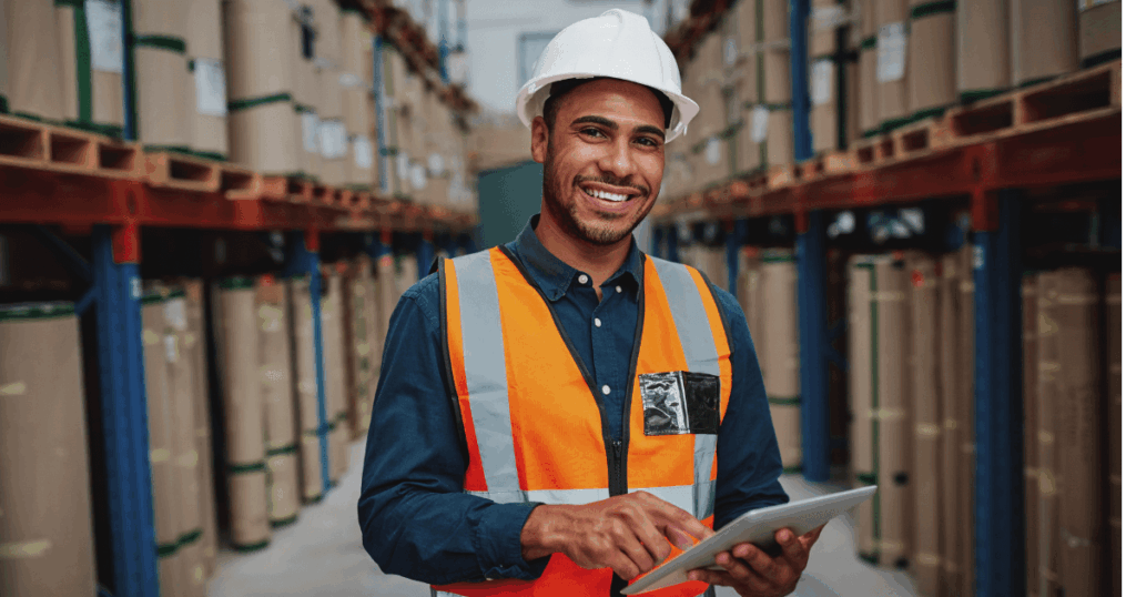Profissional em um armazém, usando colete de segurança e capacete, operando um tablet enquanto gerencia o estoque, contribuindo para a prevenção de perdas e controle eficiente de mercadorias.