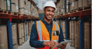 Profissional em um armazém, usando colete de segurança e capacete, operando um tablet enquanto gerencia o estoque, contribuindo para a prevenção de perdas e controle eficiente de mercadorias.