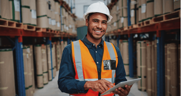 Profissional em um armazém, usando colete de segurança e capacete, operando um tablet enquanto gerencia o estoque, contribuindo para a prevenção de perdas e controle eficiente de mercadorias.