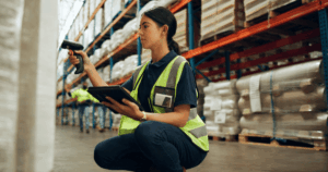 Funcionária de armazém escaneando produtos com leitor de código de barras e tablet, monitorando o inventário para evitar a ruptura de estoque.