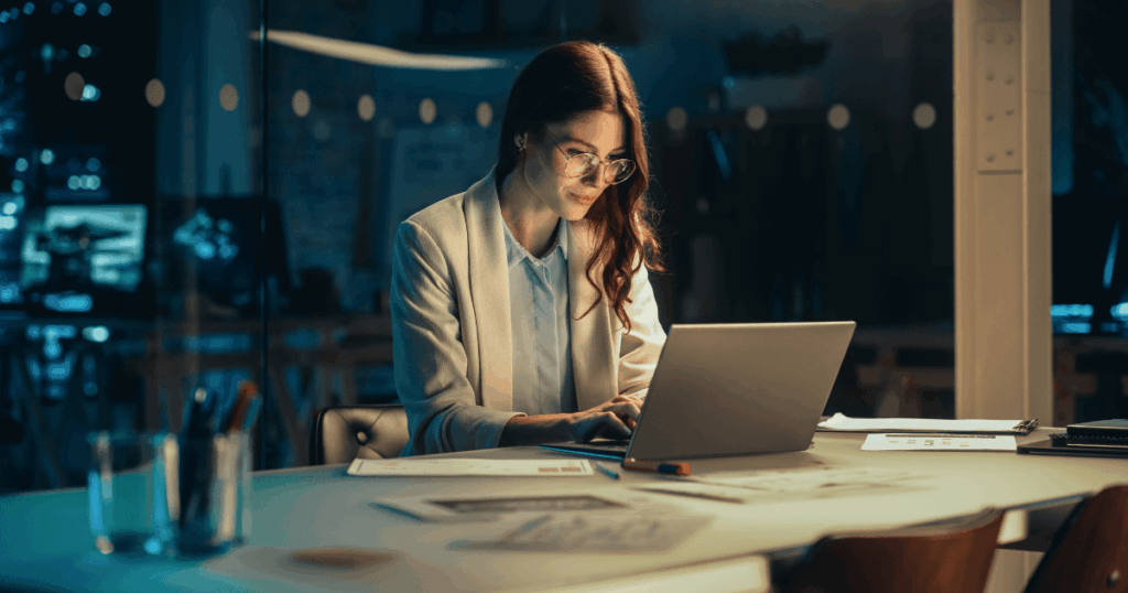 Mulher trabalhando em um escritório à noite diante do laptop, analisando documentos e realizando atividades relacionadas ao adicional noturno.