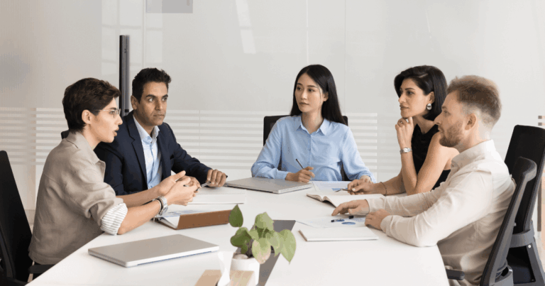 Reunião de um comitê gestor da reforma tributária, com seis pessoas sentadas ao redor de uma mesa discutindo documentos e análises em um ambiente de escritório moderno.