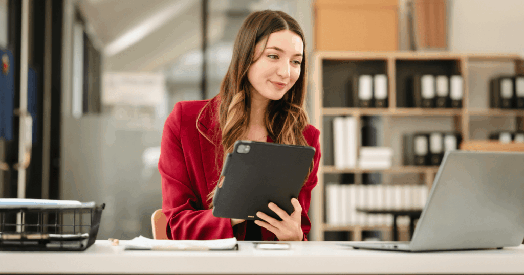 Mulher sentada em um escritório, segurando um tablet e olhando para um notebook, representando atividades de compliance fiscal e análise de conformidade tributária.