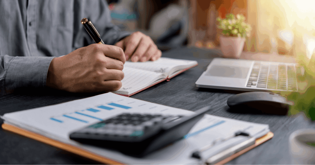 Pessoa trabalhando em uma mesa de escritório, fazendo anotações em um caderno ao lado de gráficos financeiros, calculadora e laptop, ilustrando atividades de análise e gestão relacionadas ao IBS.