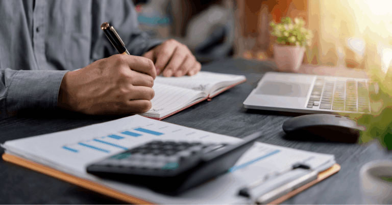 Pessoa trabalhando em uma mesa de escritório, fazendo anotações em um caderno ao lado de gráficos financeiros, calculadora e laptop, ilustrando atividades de análise e gestão relacionadas ao IBS.