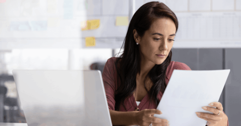 Mulher analisando documentos financeiros em frente ao computador, possivelmente revisando relatórios de ativo imobilizado em um ambiente de escritório.