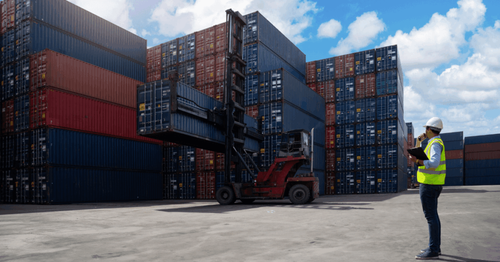 Pátio de contêineres com empilhamento de cargas, empilhadeira movimentando um contêiner e trabalhador com EPI conferindo documentos, ilustrando operações de comércio exterior e drawback na logística portuária