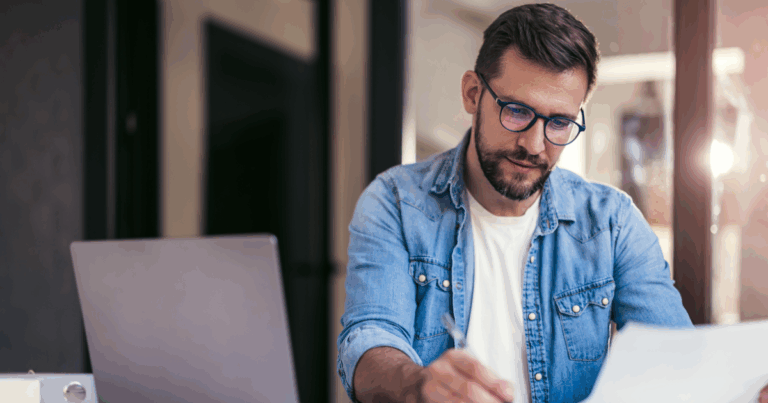 Homem usando óculos trabalha concentrado em uma mesa com notebook e documentos em um ambiente de escritório, representando análise profissional relacionada a IBS e CBS.