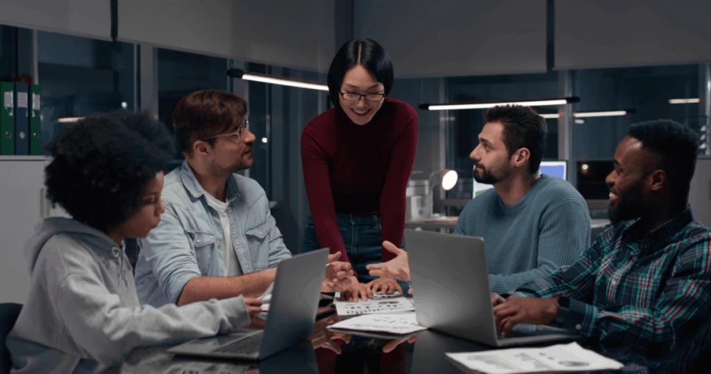 Equipe diversa reunida em escritório colaborativo discutindo um projeto de inovação com laptops e documentos sobre a mesa.