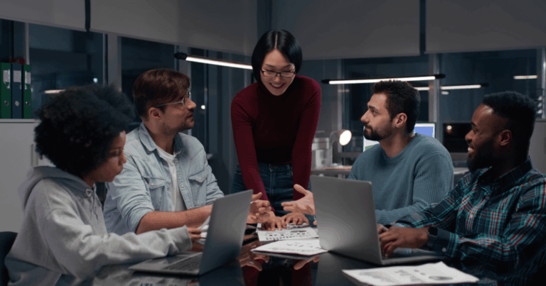 Equipe diversa reunida em escritório colaborativo discutindo um projeto de inovação com laptops e documentos sobre a mesa.