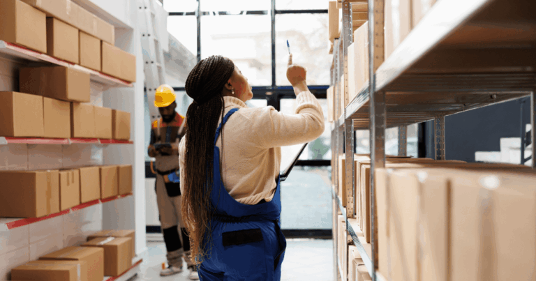Funcionária realizando a separação de pedidos em um armazém, conferindo caixas nas prateleiras enquanto outro trabalhador organiza itens ao fundo.