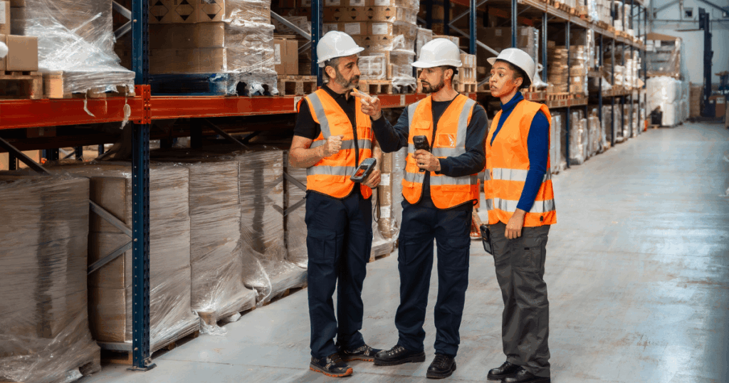 Profissionais com equipamentos de segurança conversando em um armazém logístico, analisando processos da cadeia de abastecimento entre estoque, armazenagem e distribuição de mercadorias.