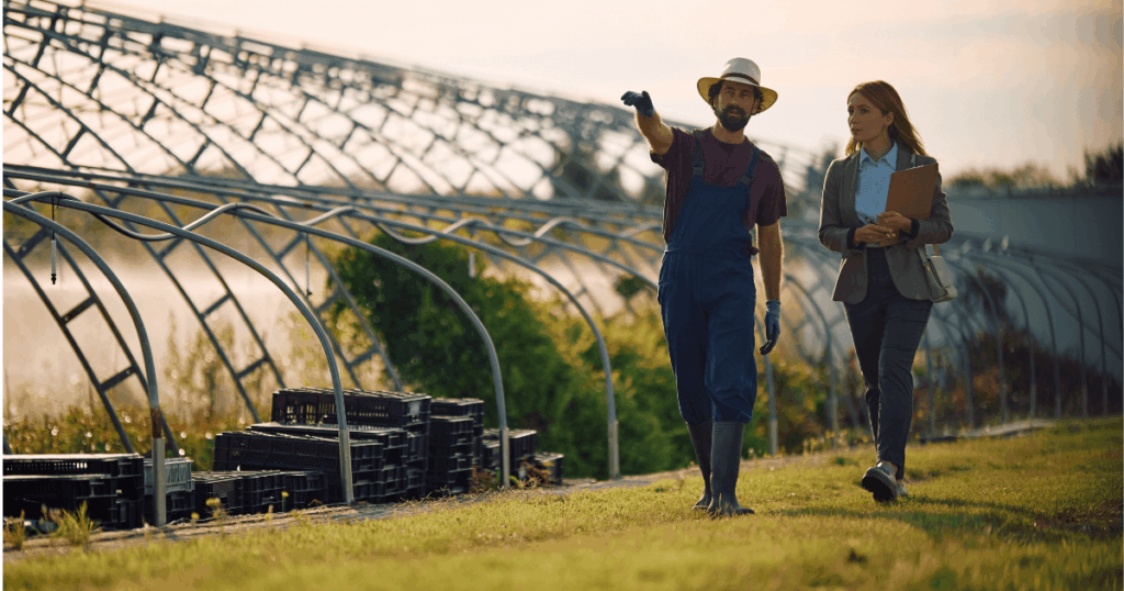 Profissional rural aponta estruturas de estufa enquanto conversa com consultora com prancheta, representando práticas de gestão agrícola no planejamento e monitoramento da produção.