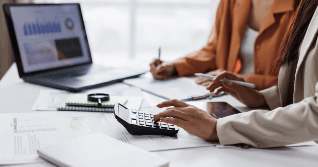 Profissionais analisando relatórios financeiros e utilizando calculadora sobre a mesa, com gráficos no notebook, ilustrando impacto da taxa Selic em análises econômicas e financeiras.