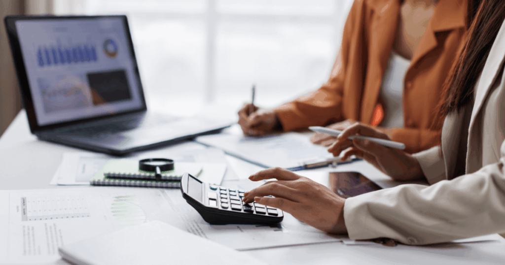 Profissionais analisando documentos financeiros com calculadora e notebook em mesa de escritório, representando atividades de contabilidade empresarial.