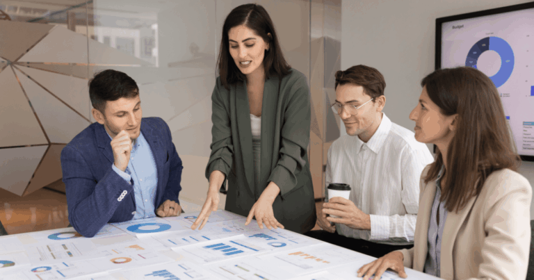 Equipe de profissionais reunida em escritório analisando gráficos e relatórios sobre a mesa, discutindo dados estratégicos em uma análise de mercado.