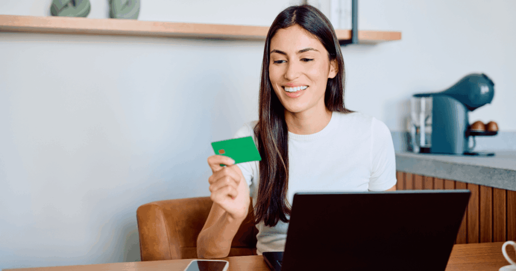 Mulher sorrindo enquanto segura um cartão de crédito e usa o notebook para fazer uma compra online, representando o comportamento do consumidor no ambiente digital.