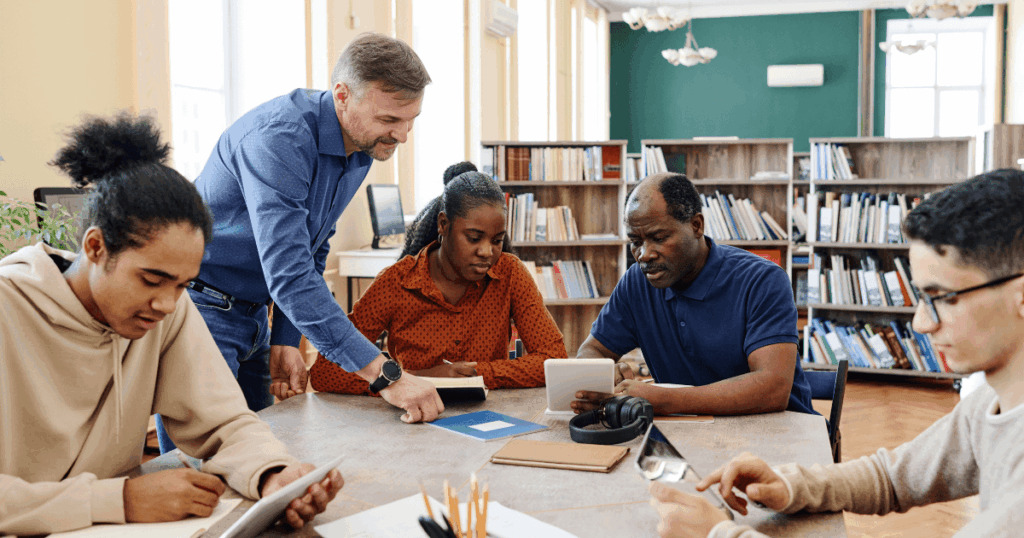 Grupo diverso de pessoas em uma biblioteca, com um instrutor orientando enquanto todos utilizam tablets e fazem anotações, representando colaboração e lifelong learning em um ambiente de estudo contínuo.