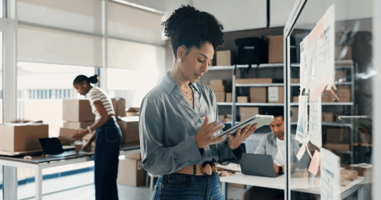 Profissionais trabalhando em um escritório de e-commerce, utilizando tablet e notebook para gerenciar pedidos, logística e operações em um ambiente de armazenagem com caixas e materiais de envio.
