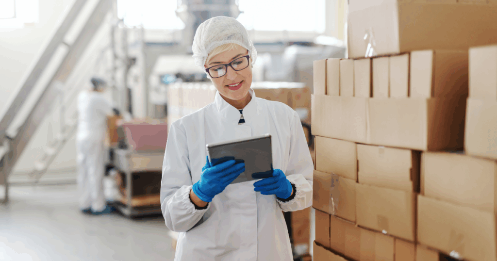 Profissional da indústria alimentícia, usando uniforme de proteção, luvas e touca, confere informações em um tablet ao lado de caixas empilhadas em um centro de produção, ilustrando o processo de rastreabilidade de alimentos para controle de qualidade, segurança e gestão de estoque.