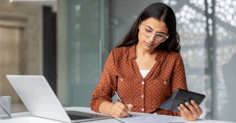 Mulher analisando documentos e fazendo anotações enquanto usa um tablet e um notebook, representando atividades de cálculo e gestão tributária relacionadas ao DIFAL